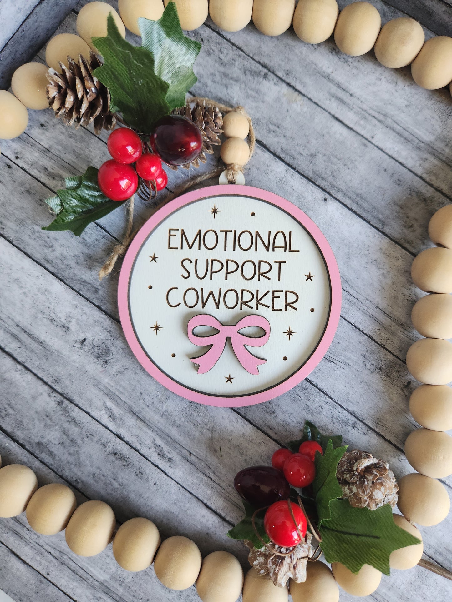 Decorative sign with 'Emotional Support Colleague' text and pink ribbon on a wooden background with beads and holly.