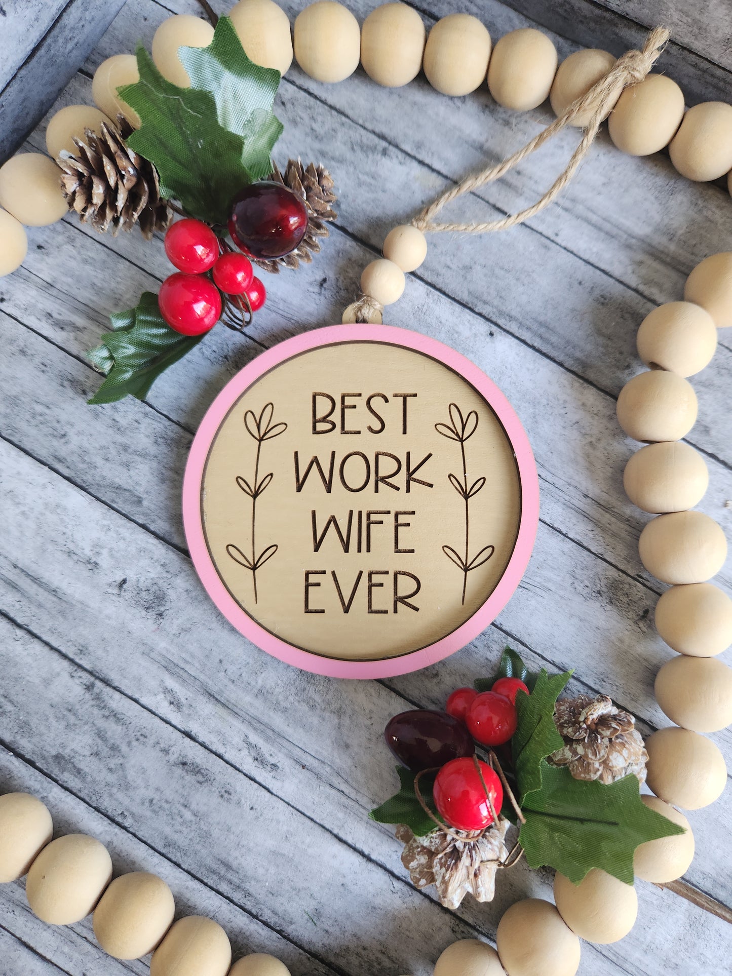 Decorative round sign with 'Best Work Wife Ever' text, surrounded by wooden beads and holly leaves on a wooden surface.