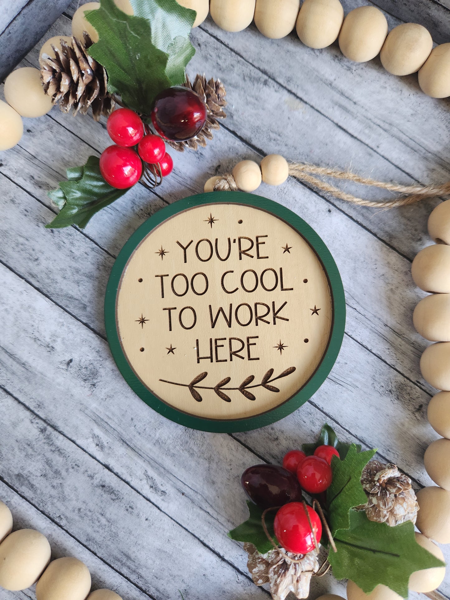 Decorative sign with text 'You're too cool to work here' surrounded by Christmas decorations on a wooden surface.