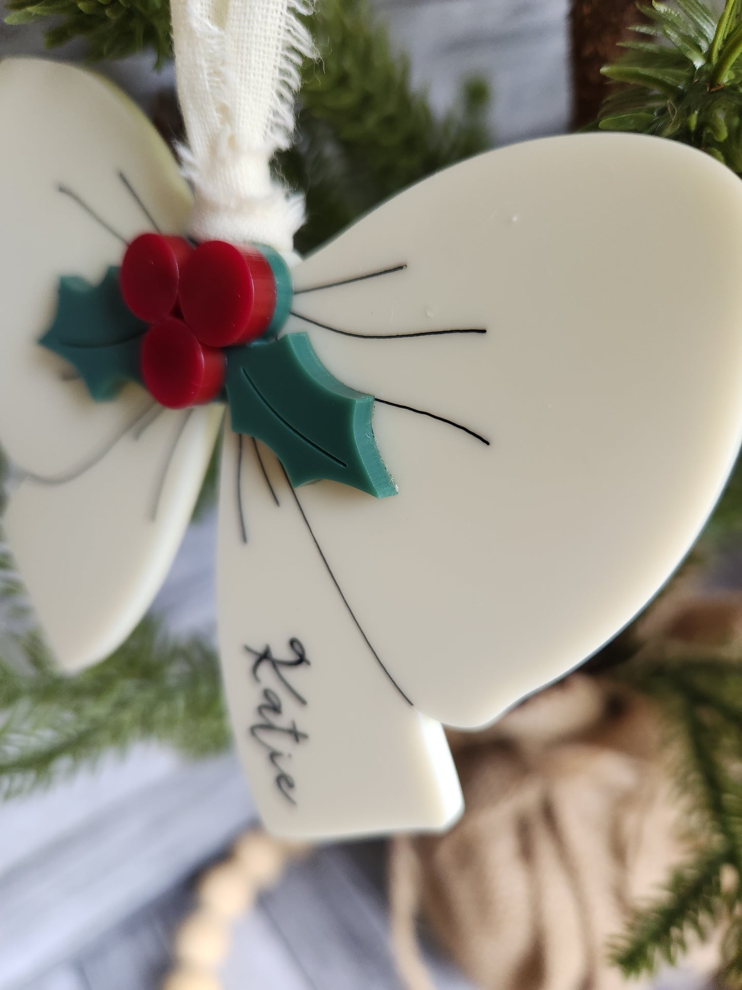 Decorative angel ornament with holly design and 'Katie' inscription on a blurred Christmas tree background.