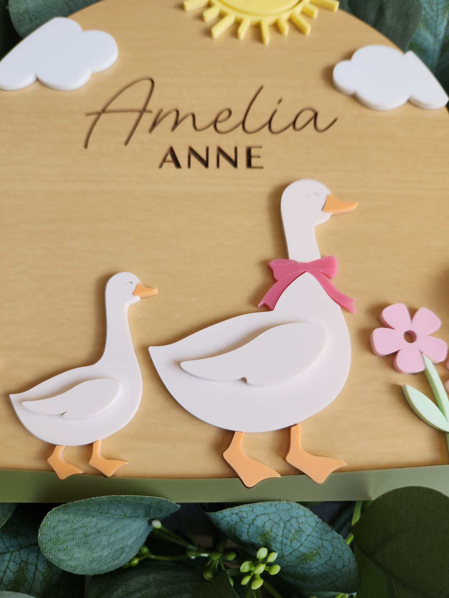 Wooden plaque with decorative elements and 'Amelia Anne' engraving, surrounded by greenery.