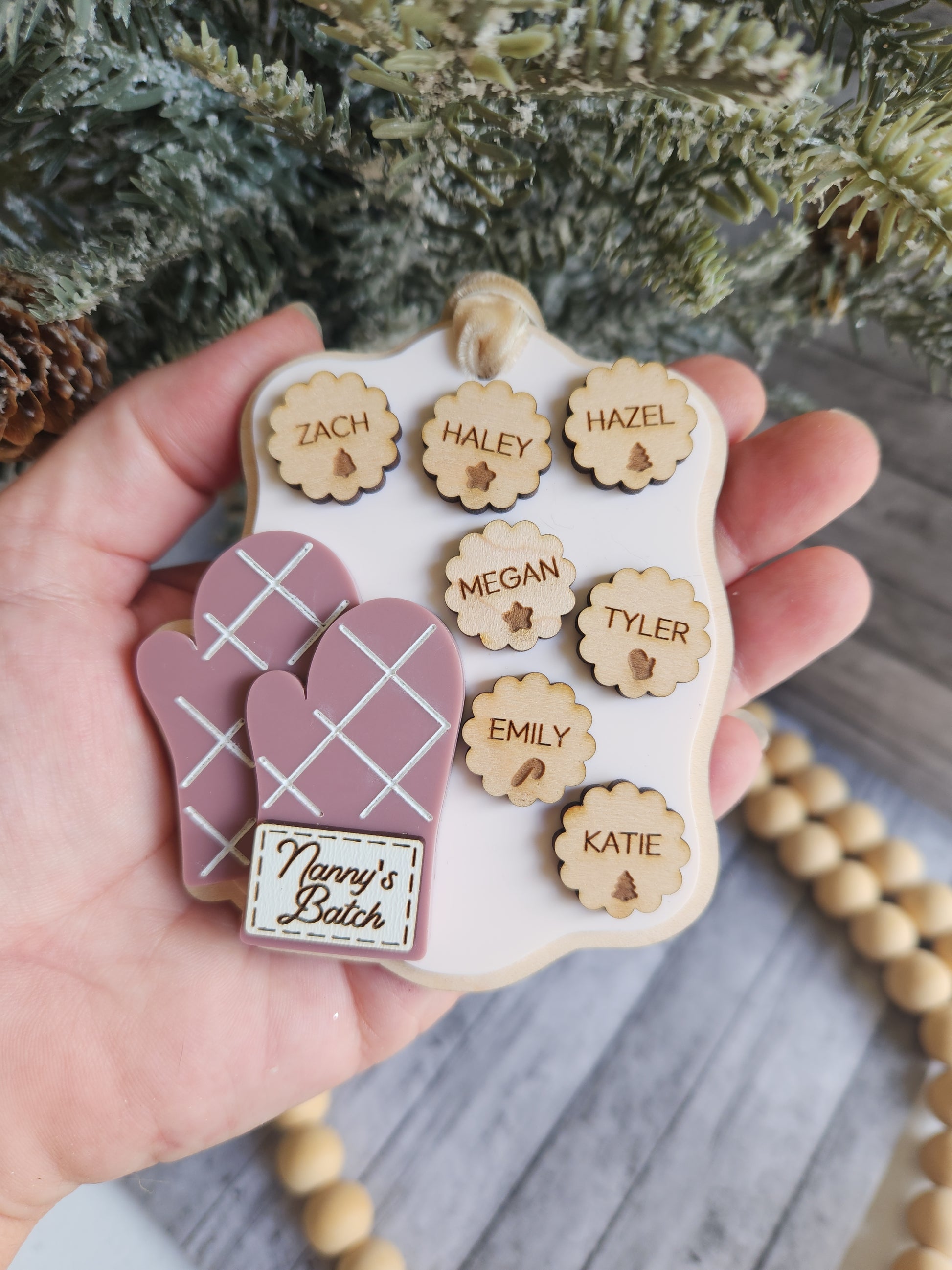 Hand holding a wooden name plate with engraved names and a pink heart-shaped item, with a blurred background.