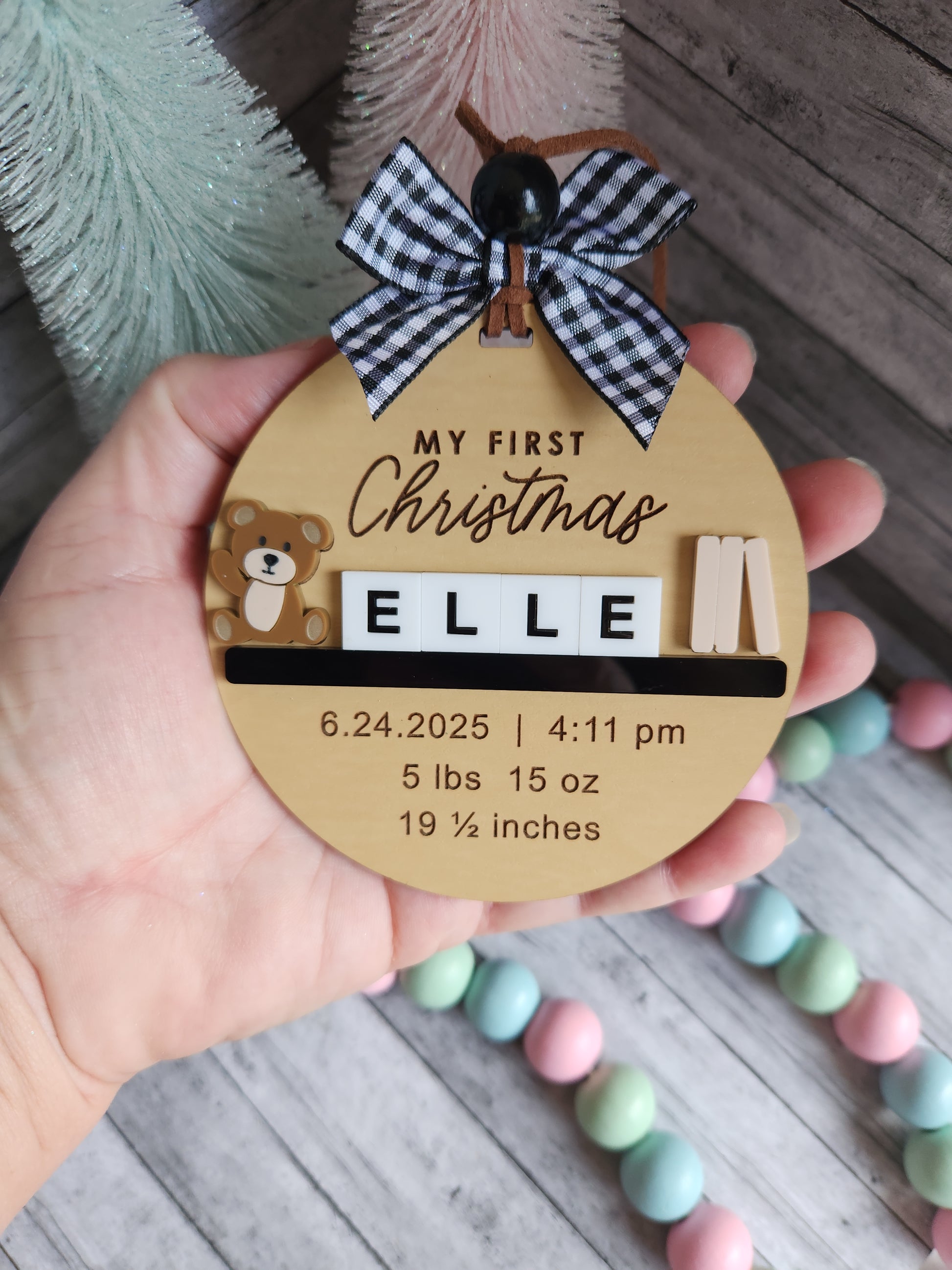 Round wooden tag with baby's first Christmas details held by a hand, with colorful beads in the background.