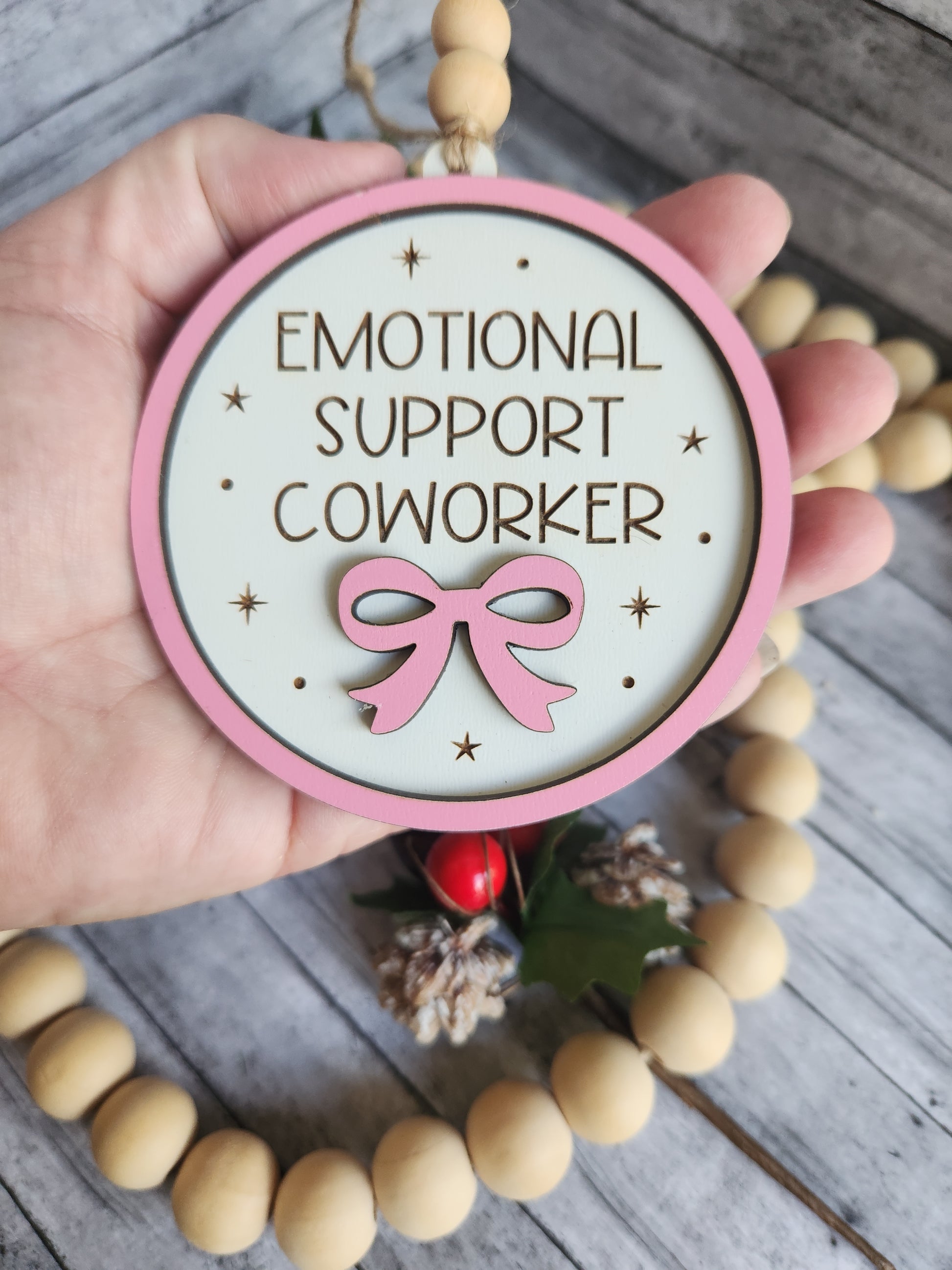 Decorative sign with 'Emotional Support Coworker' text and pink ribbon held by a hand on a wooden surface.