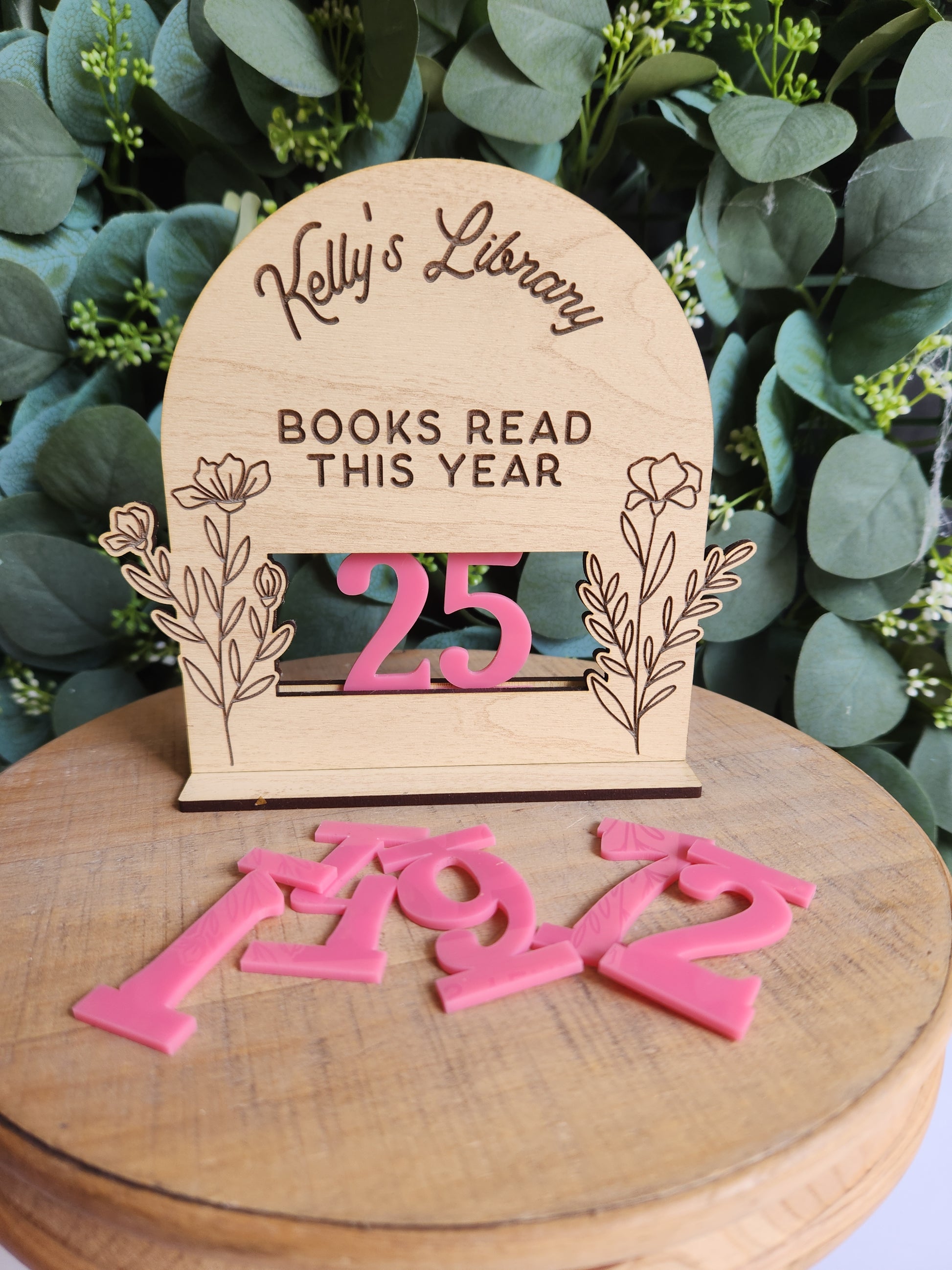 Wooden book marker with 'Kelly's Library' and '25 books read this year' text, surrounded by greenery.
