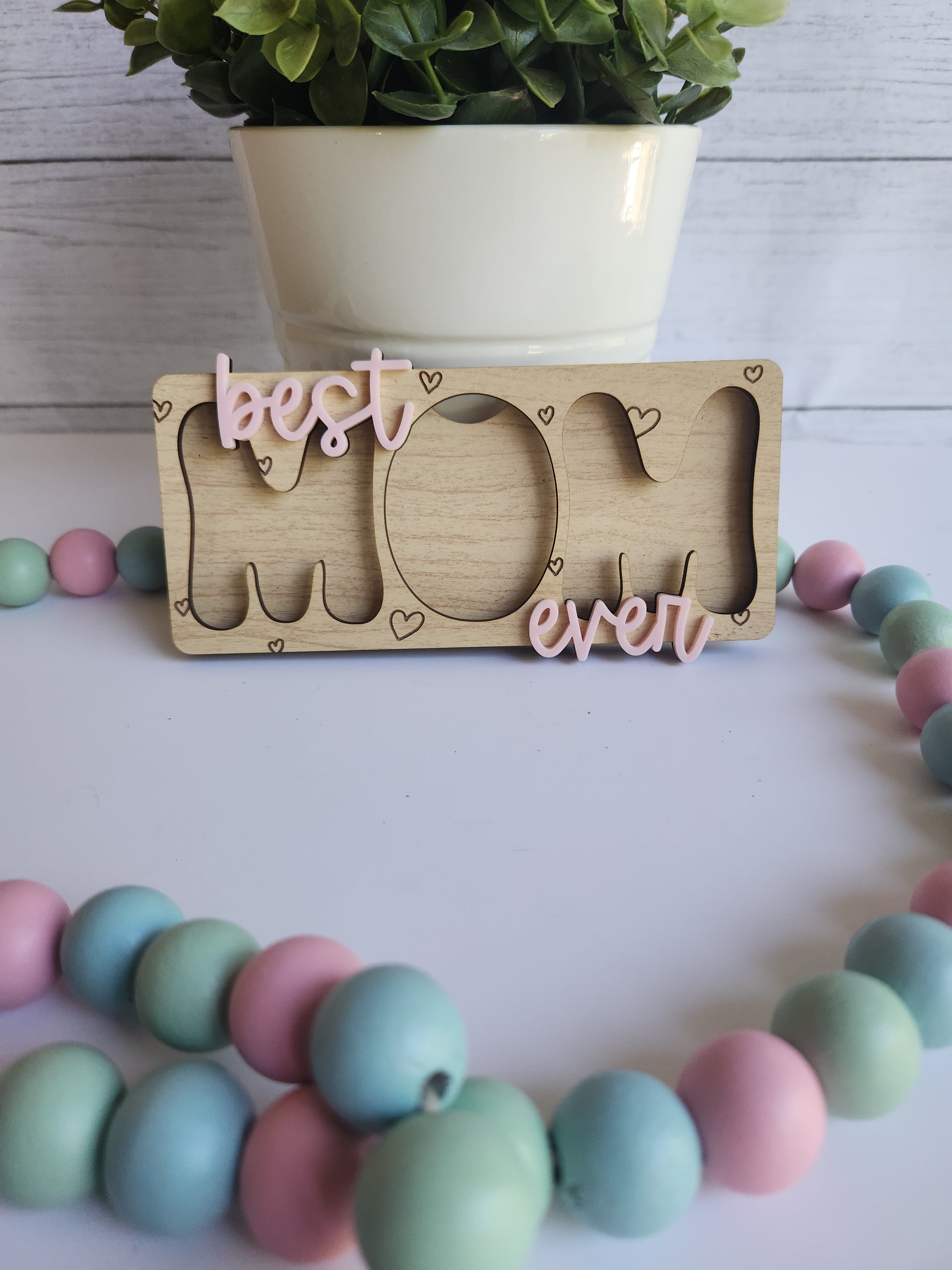Wooden card with cut-out shapes and 'best ever' text, next to a colorful beaded necklace on a light wooden surface.