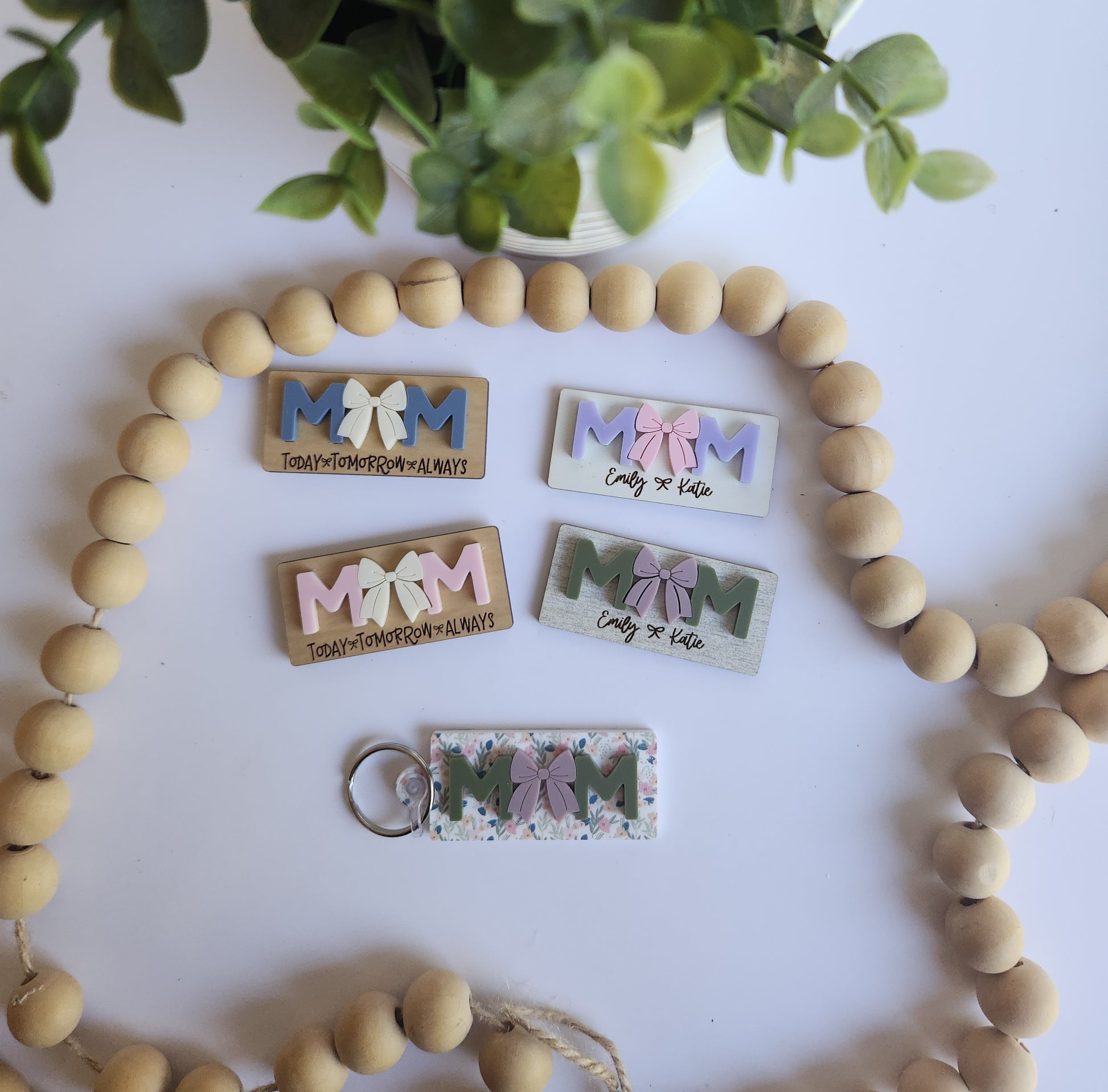 A collection of personalized 3D MOM magnets and keychains in various colors like sage green, dusty blue, and floral patterns. The designs feature layered acrylic letters with a center bow and custom engraved names like 'Emily & Katie' or the phrase 'Today, Tomorrow, Always' on wood and acrylic bases