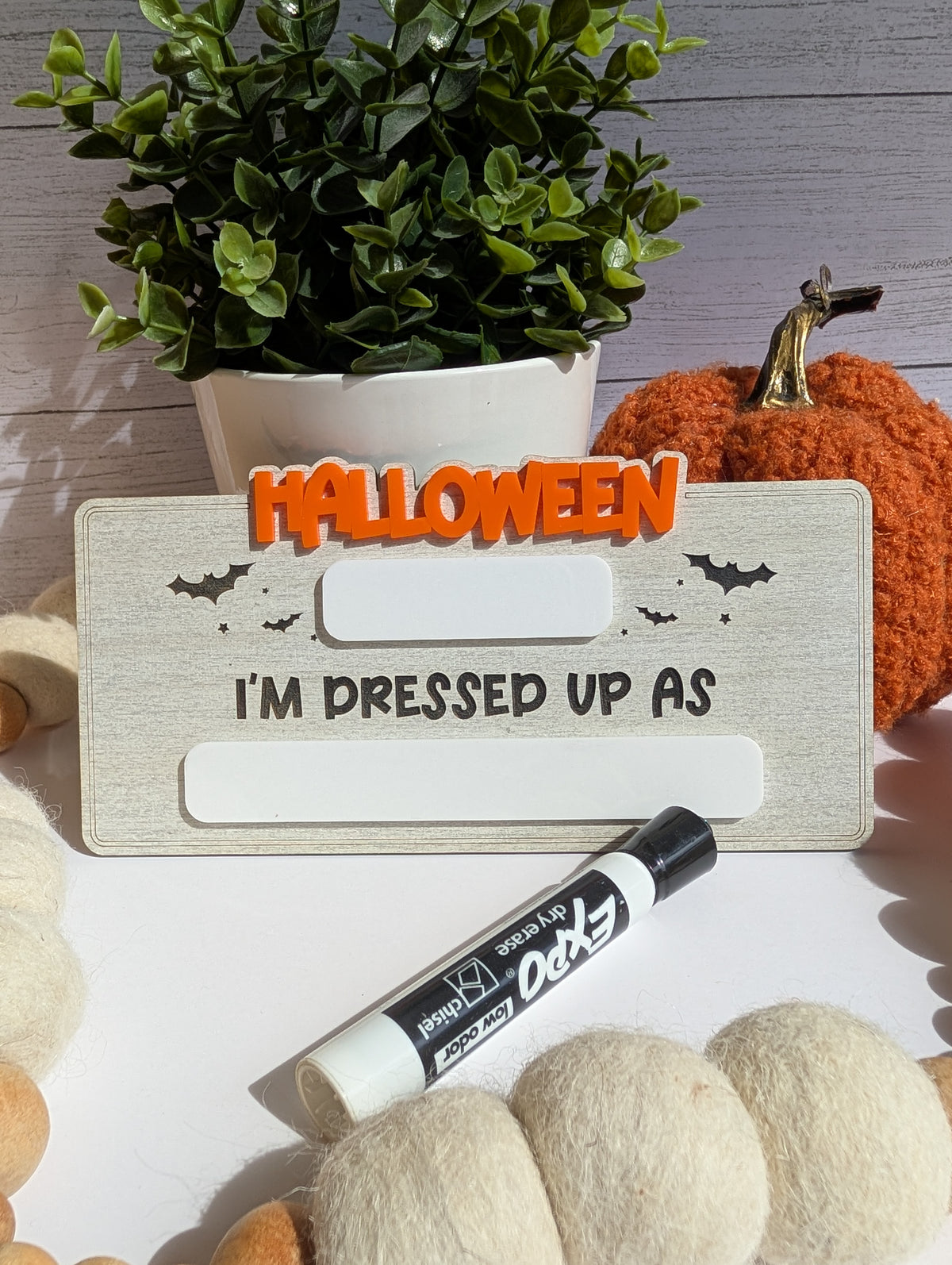 Reusable wooden Halloween costume photo prop sign in whitewashed wood with raised orange Halloween lettering, black bat accents, and two blank dry-erase sections for writing the year and child's costume.